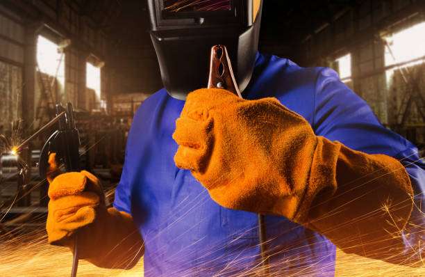 Welder in mask and orange welding glove holds tools with sparks flying in industrial setting.